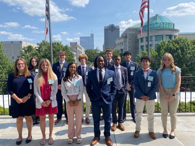 12 Senate Pages posing on a sunny day at the legislative building in front of flag poles and museums